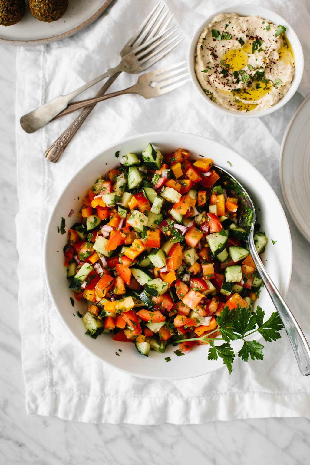Israeli salad next to a small bowl of baba ganoush and falafel. Israeli salad made from tomatoes, cucumber, bell pepper, red onion and herbs in a white bowl.