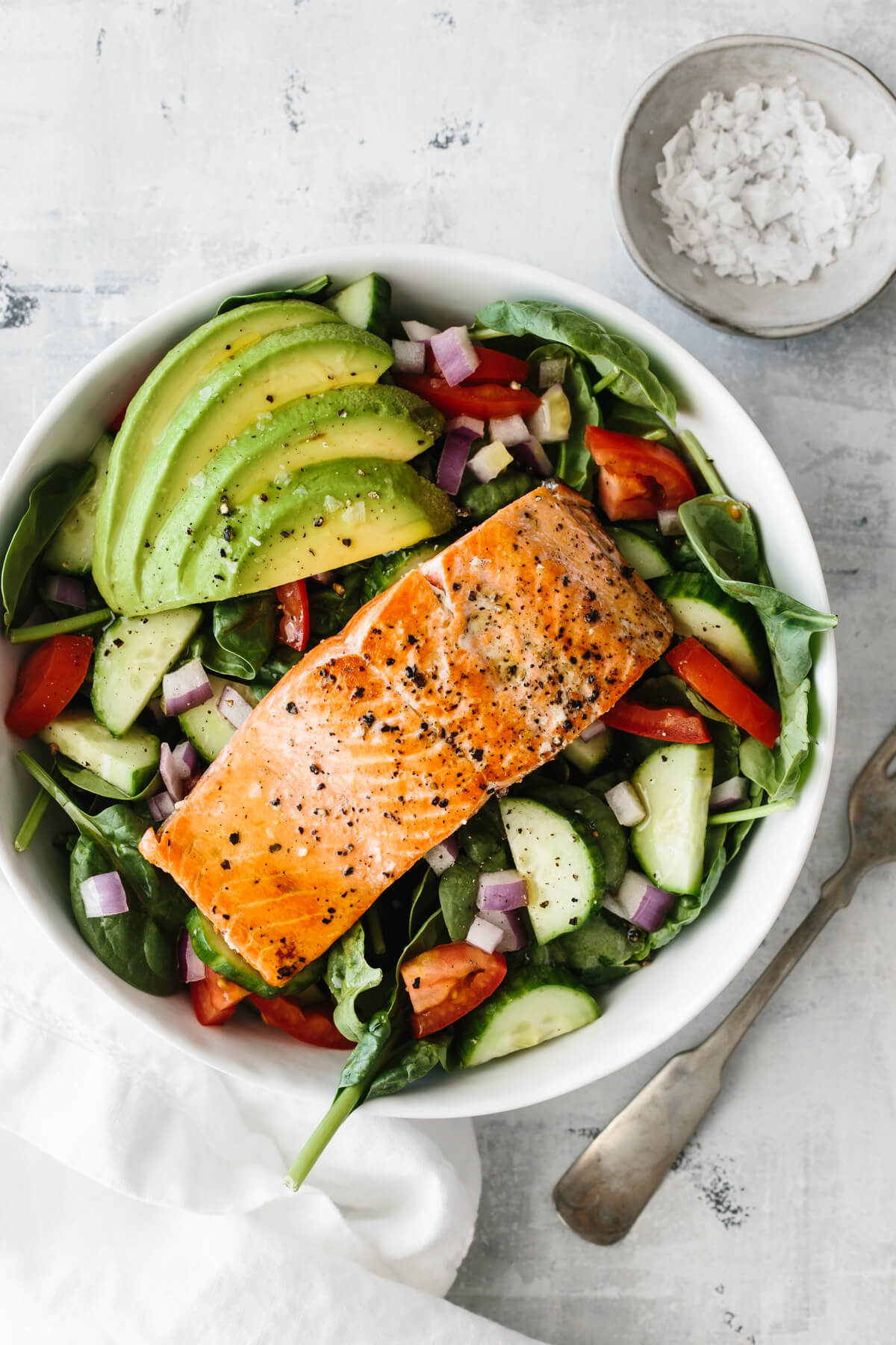 Single salmon fillet on top of salad with avocado and salt pinch bowl off to the side.