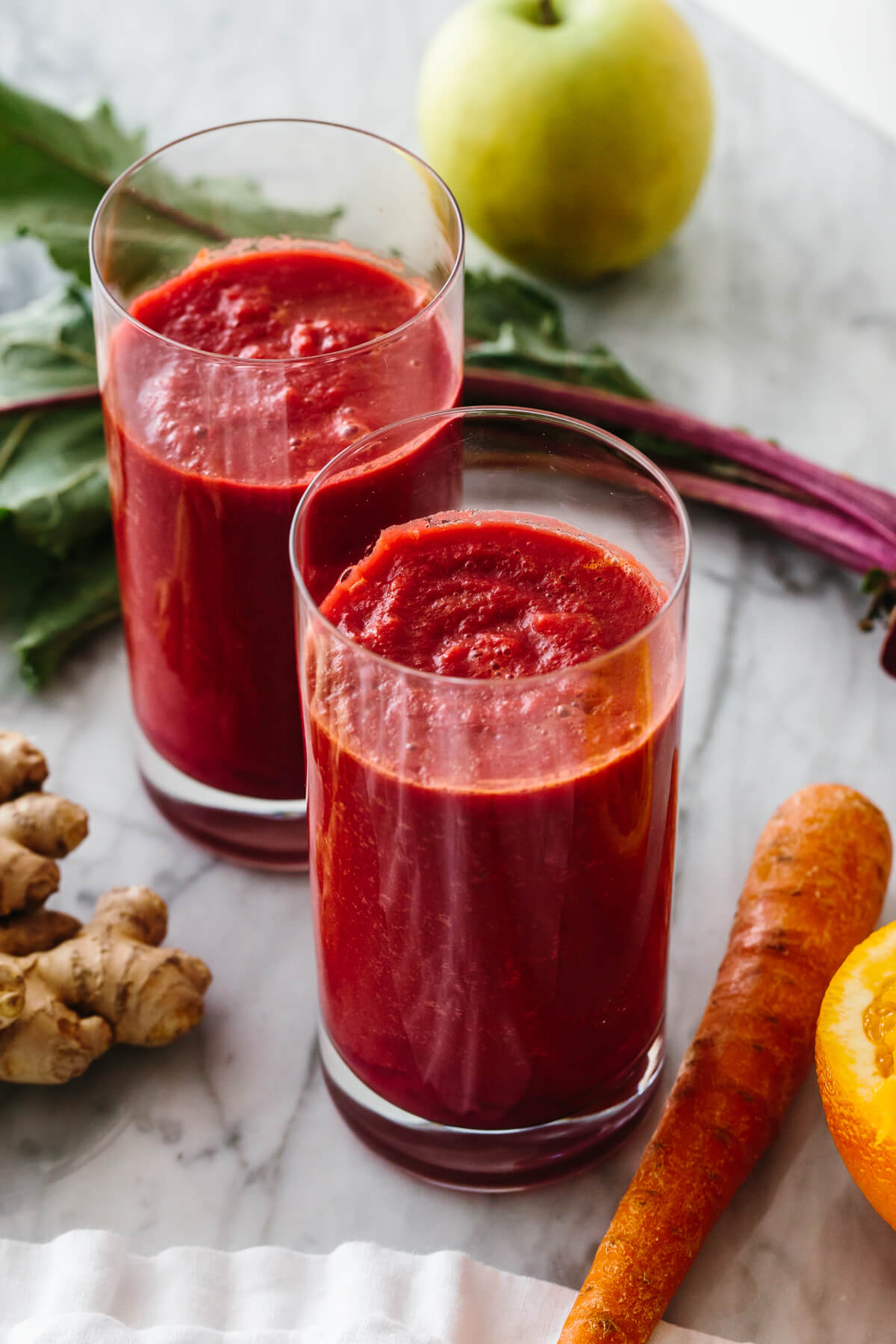 Two tall glasses of smoothie, surrounded by carrots, beets, apple and ginger.