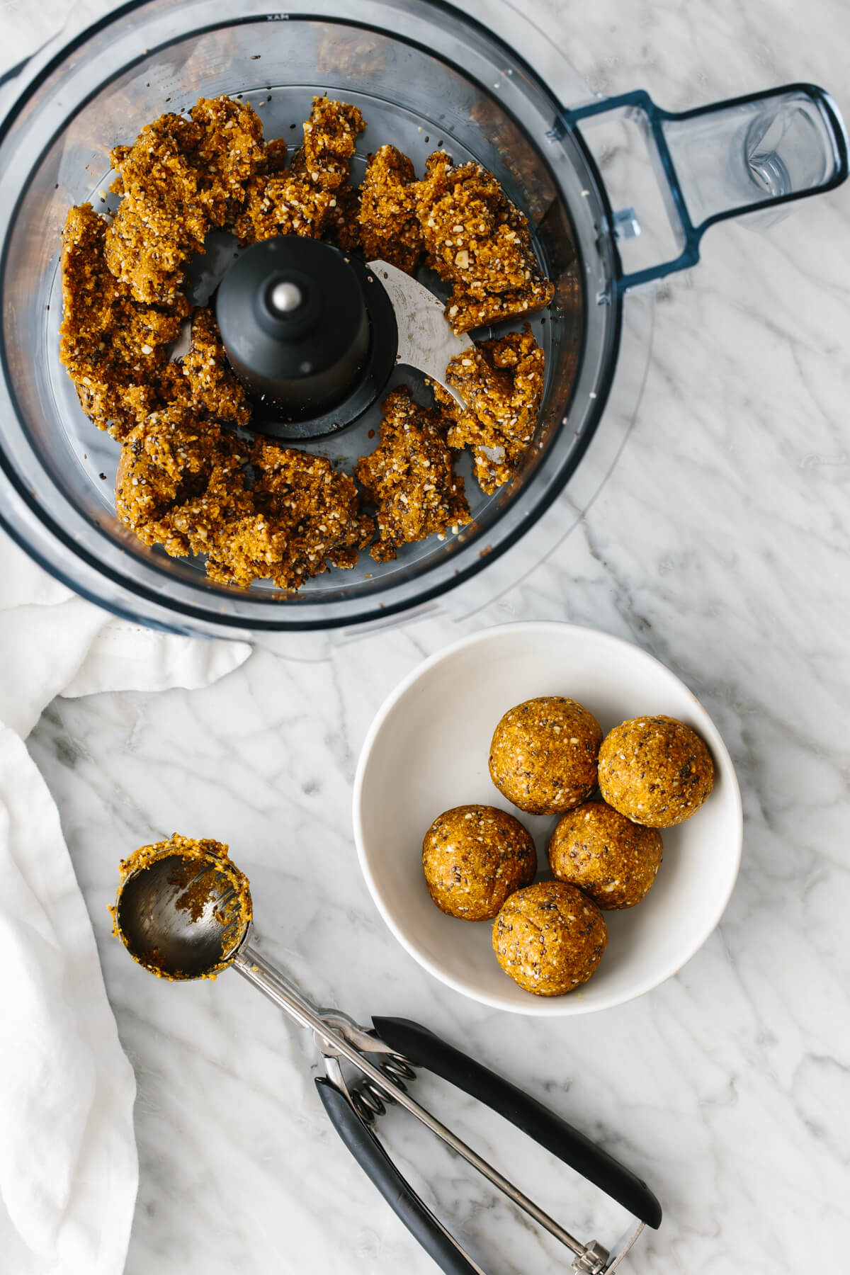 A food processor with blended mango date energy ball dough inside next to a white bowl.