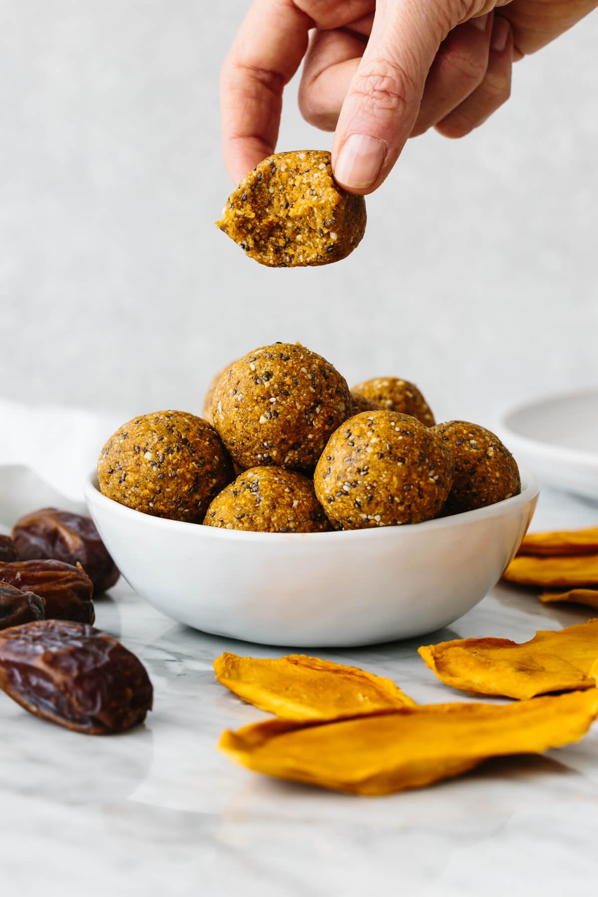 Hand picking up a bitten mango energy ball above a bowl of them.