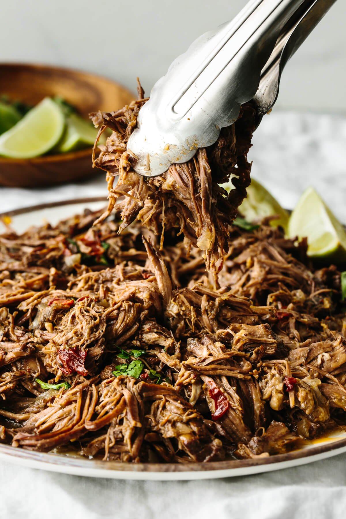Mexican beef barbacoa that's juicy, meltingly tender, seasoned with flavorful chilies and spices, and made easy right in the slow cooker! Picking up beef barbacoa from a plate with tongs.