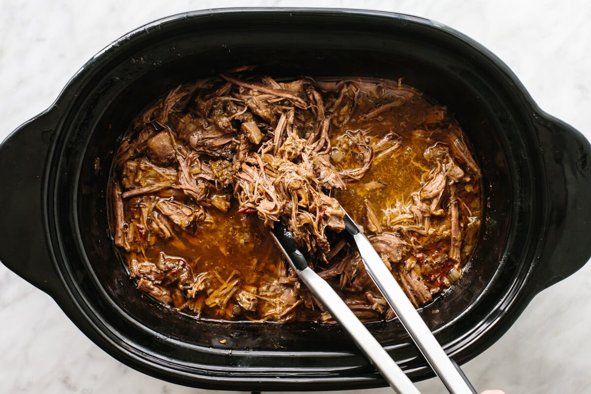 Mexican beef barbacoa that's juicy, meltingly tender, seasoned with flavorful chilies and spices, and made easy right in the slow cooker! Picking up beef barbacoa in a slow cooker with tongs.