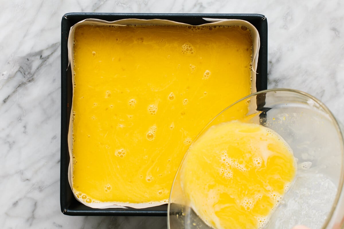 These lemon bars have a gluten-free shortbread-like crust and a bright, luscious filling that's the perfect balance of sweet and tangy! Pouring lemon bar filling into a pan with the crust.