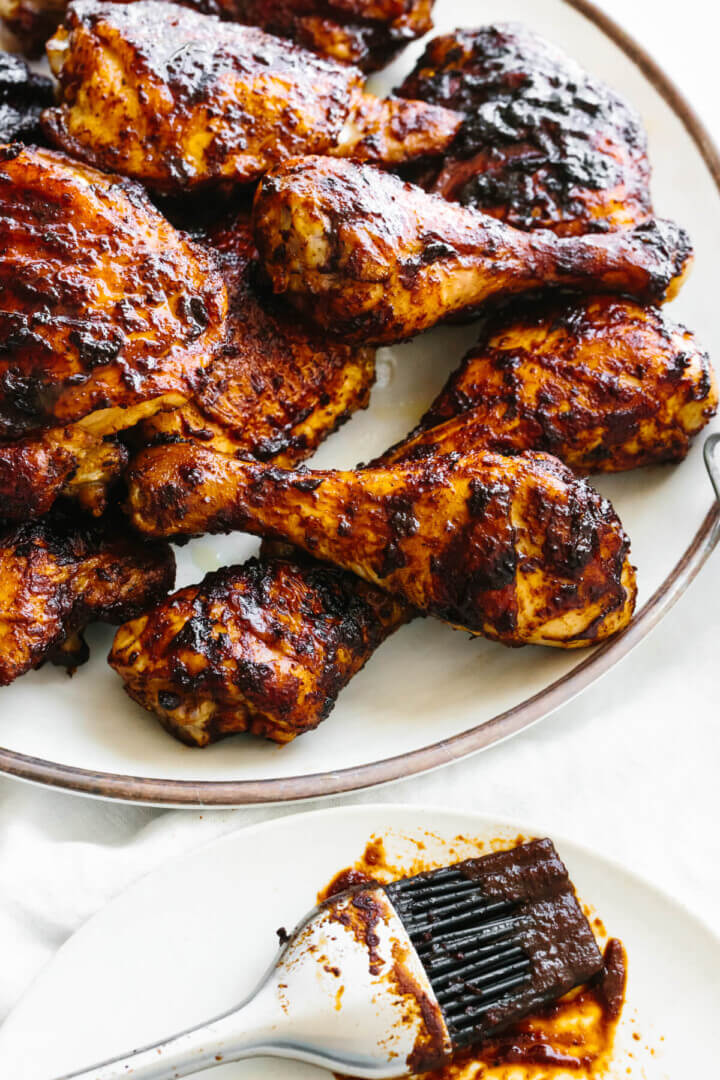 This BBQ chicken is insanely juicy, perfectly charred, and glistening with my secret homemade BBQ sauce. Perfect for your summer cookout! A white plate of BBQ chicken thighs and drumsticks