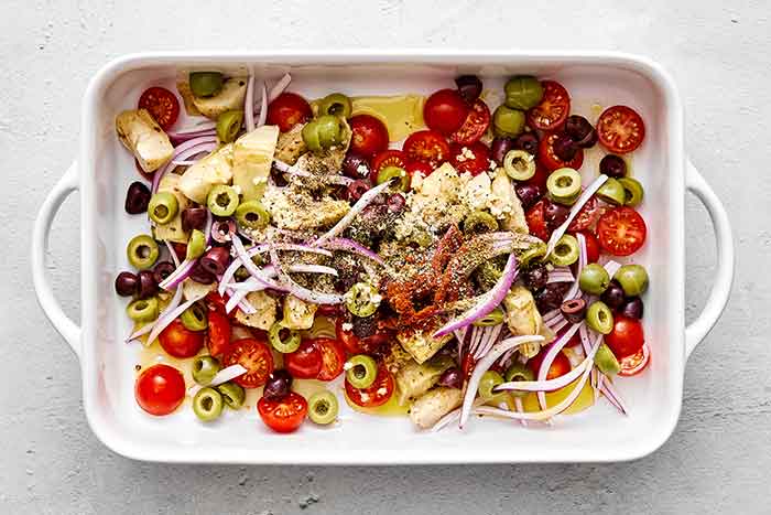 Mixing Greek veggies with seasoning in a casserole pan.