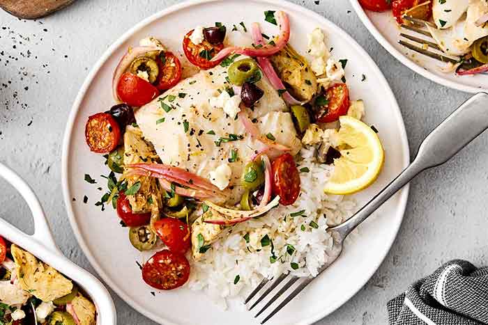 Greek baked cod on a plate with veggies and rice.