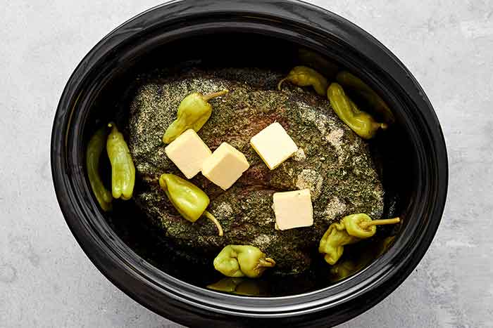 Adding butter and banana peppers to chuck roast in a crockpot.